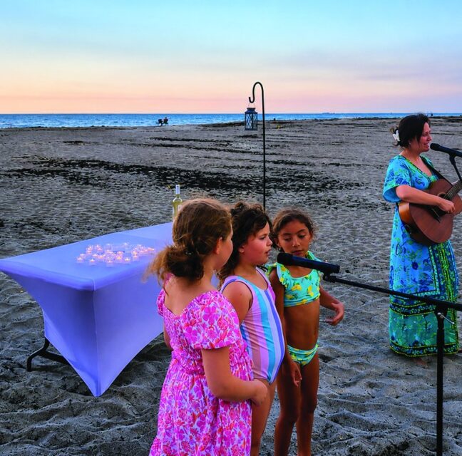 Children gather as Cantor Abbie Strauss leads Beach Shabbat with guitar at sunset
