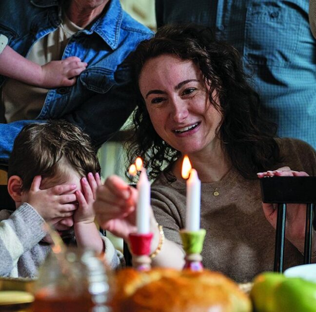 Mother lights Shabbat candles while her young son covers his eyes