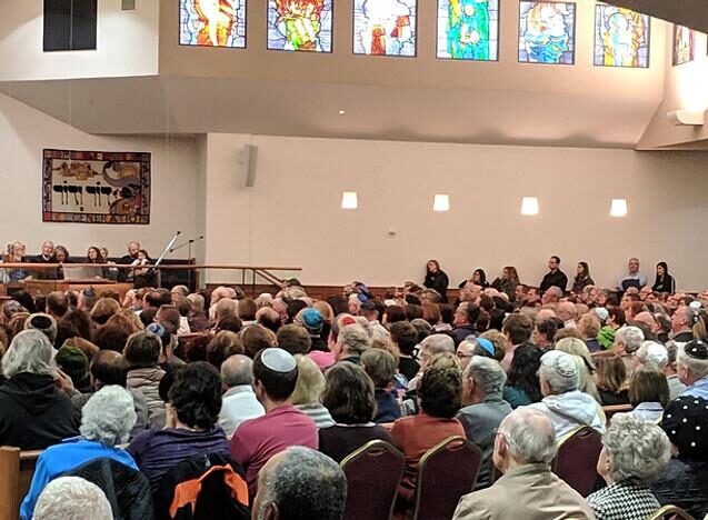 Worshippers in kippot fill a synagogue sanctuary beneath stained-glass windows